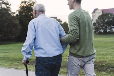 A younger man supports an elderly man with a cane while walking outdoors.
